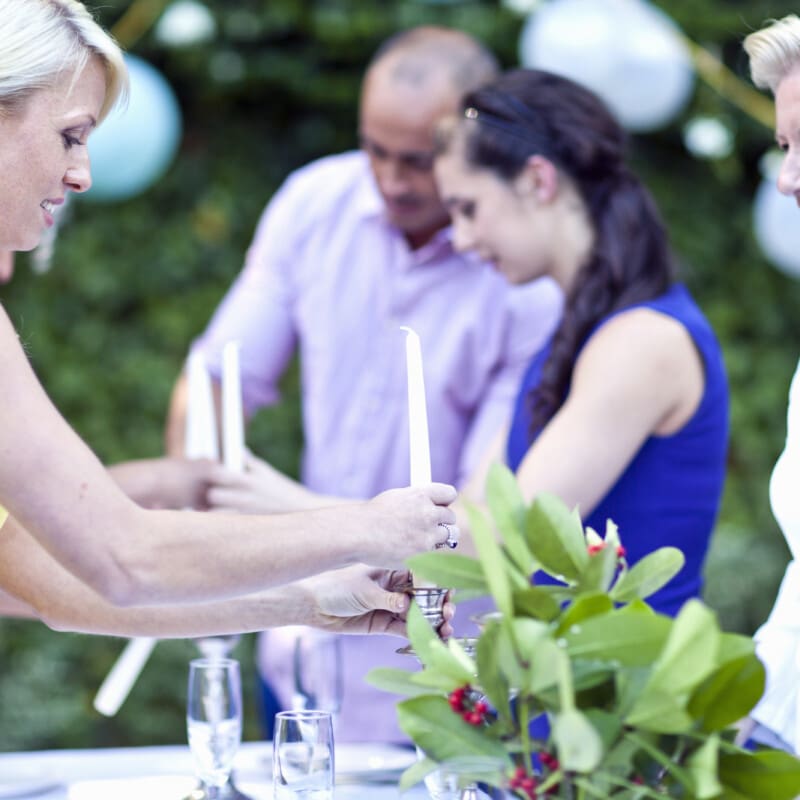 A group of people gather outdoors around a table setting up for an event. A woman in a yellow dress lights candles, while others smile and engage in conversation.