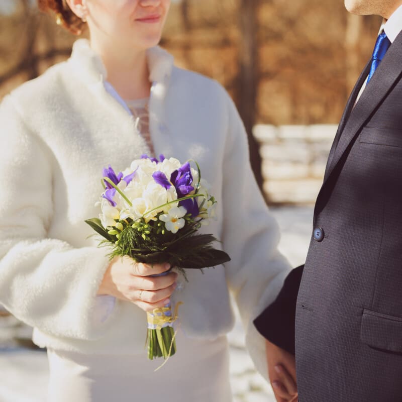 A couple stands outdoors in a winter setting; the bride in a white coat holds a bouquet of purple and white flowers, while holding hands with the groom in a dark suit.